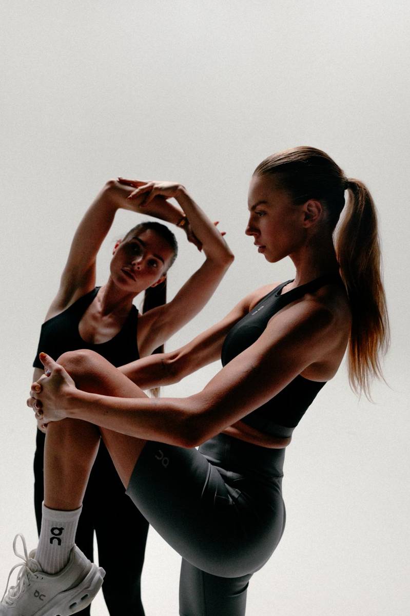 Two women training together, one guiding the other into a standing stretch